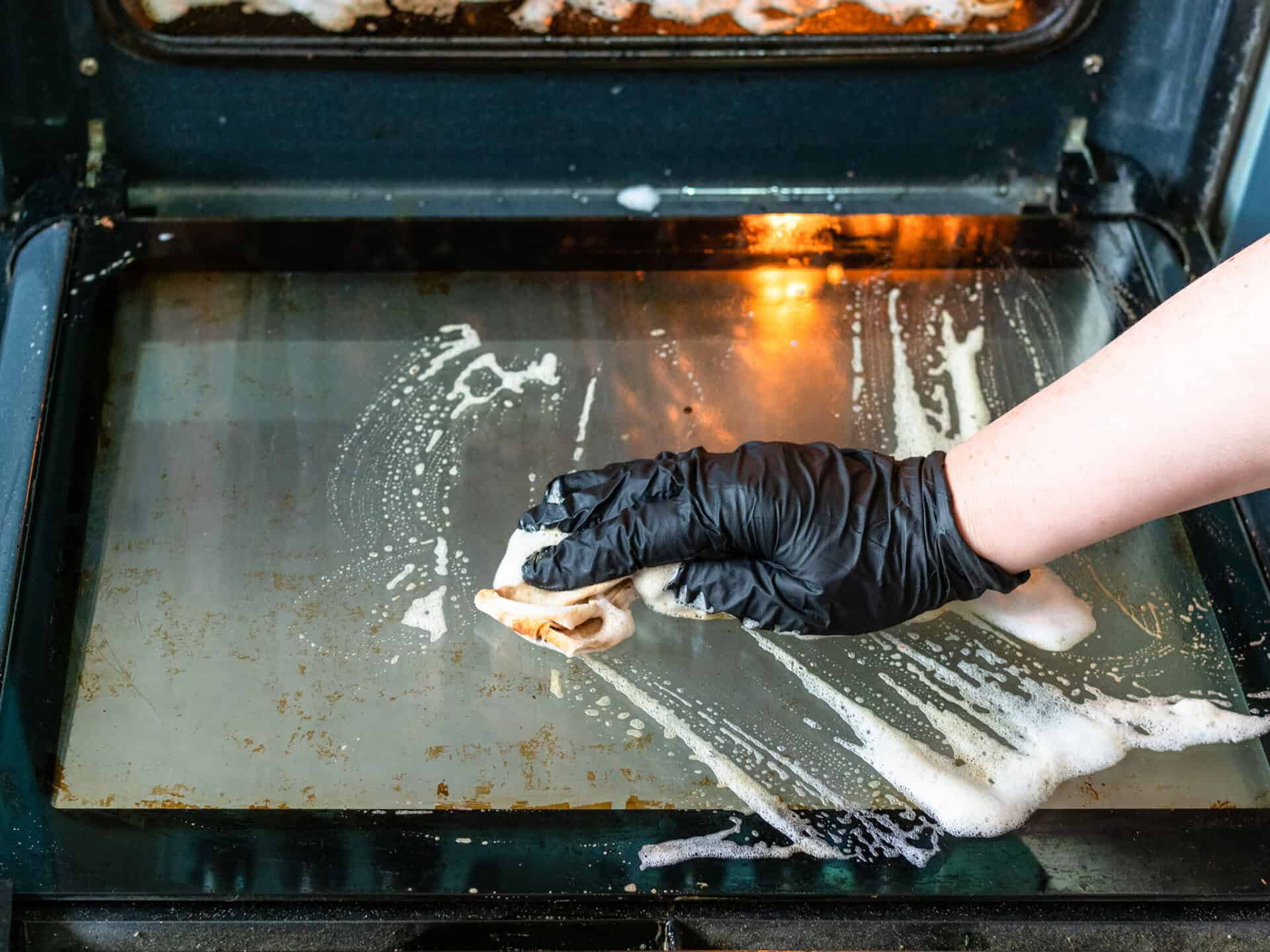 cleaning oven in home kitchen – female hand cleans glass door of dirty oven with reagent